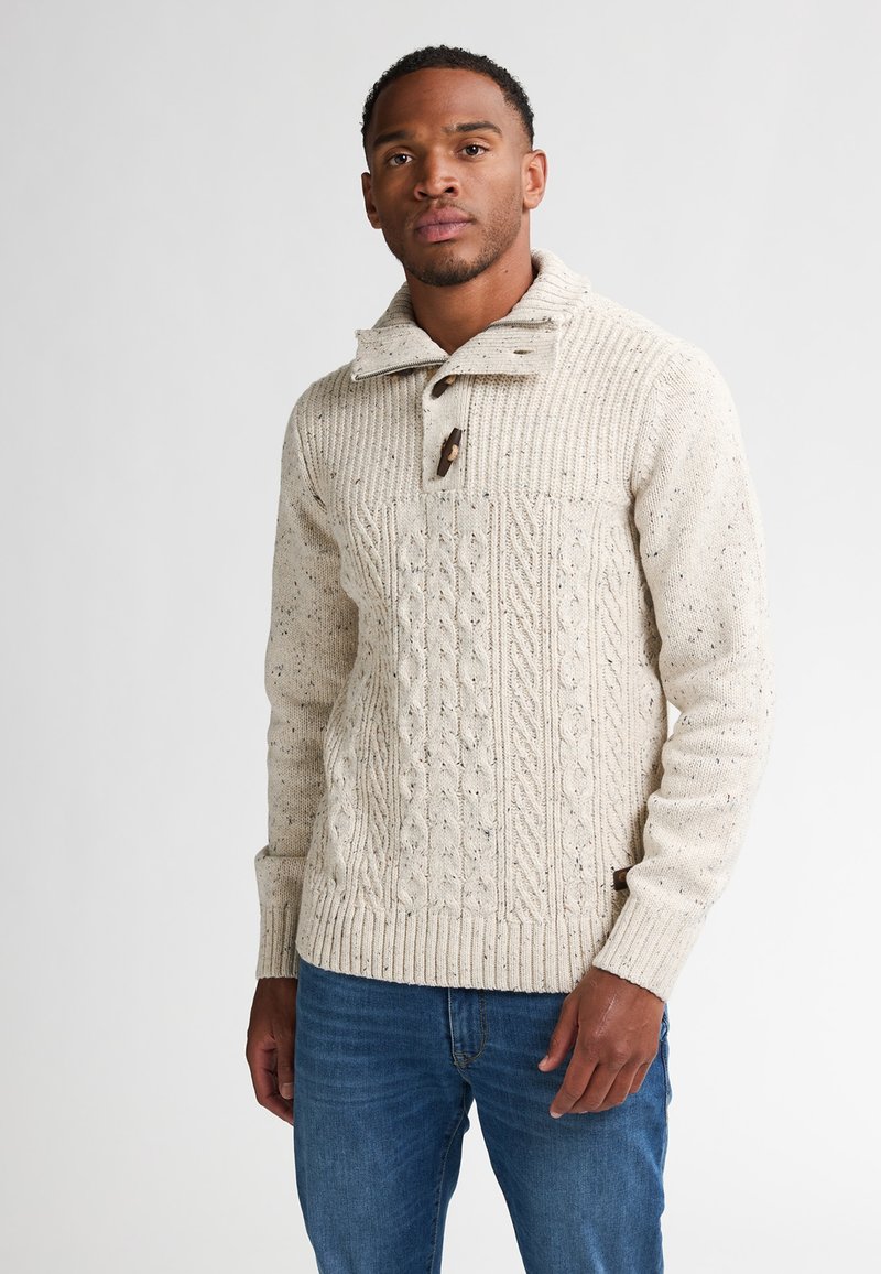 Sweter w kolorze kremowym z dzianiny w warkocze, z wysokim kołnierzem i drewnianymi guzikami, z ribbowanymi mankietami i dołem. Noszony z niebieskimi dżinsami.