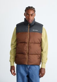 Bruine en zwarte gewatteerde vest met een hoge kraag, ritssluiting en Columbia-logo. Draagt over een groene lange mouw shirt met blauwe jeans.