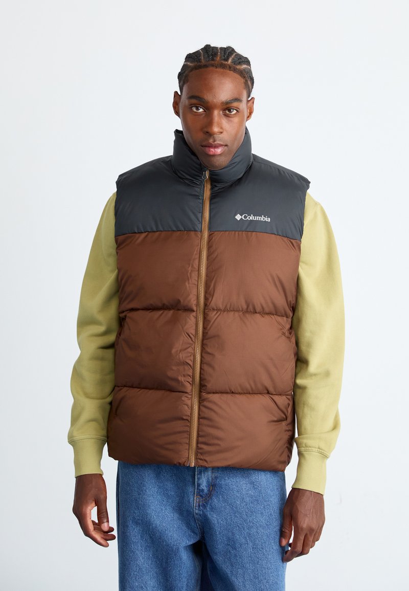 Bruine en zwarte gewatteerde vest met een hoge kraag, ritssluiting en Columbia-logo. Draagt over een groene lange mouw shirt met blauwe jeans.