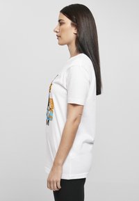 Hvit bomullst-skjorte med et fargerikt grafisk design på forsiden, korte ermer og en avslappet passform, kombinert med svarte leggings.