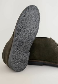 Zapato de ante verde oscuro con suela de goma negra texturizada. Presenta un diseño de cordones sencillo y bordes suaves, destacando los detalles de la artesanía.