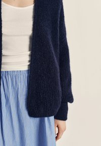 Navy blue wool cardigan with a textured finish, loose fit, and long sleeves, paired with a white tank top and blue-striped skirt.
