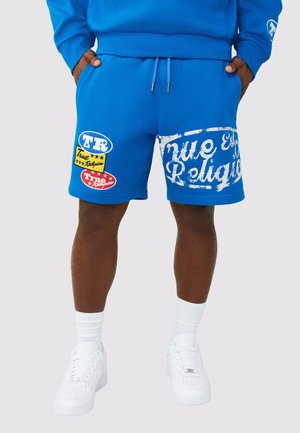 Homme portant un short bleu vif à cordon avec des logos "True Religion" et des baskets blanches, debout les mains dans les poches.