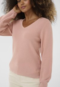 Femme aux cheveux bouclés portant un pull V rose clair et un pantalon crème, se touchant les cheveux d'une main.