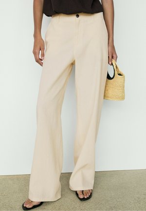 Frau trägt lockere beige Hose, schwarzes Top, schwarze Sandalen, hält kleine gewebte Strohtasche vor einfarbigem Hintergrund.