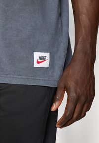 Pelēka kokvilnas T-kreklu, kuram ir balta birka ar sarkanu Nike logotipu. Apakšmalai ir neapstrādāta mala. Roku novietota tuvu krekla sāniem.