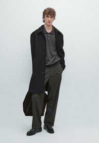 Black wool coat, gray polo shirt, pleated brown trousers, and black shoes. Brown tote bag slung over left arm; model stands straight against a plain background.