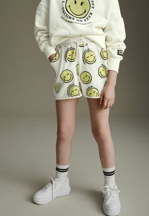 Shorts blancs avec un motif de visages souriants jaunes, taille élastique avec cordon de serrage ; associés à des baskets blanches et des chaussettes rayées.