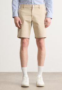 Beige bomullsshorts med brettede ermer, knapp- og glidelås-lukking, og sidelommer, kombinert med hvite sneakers og ribbede sokker.