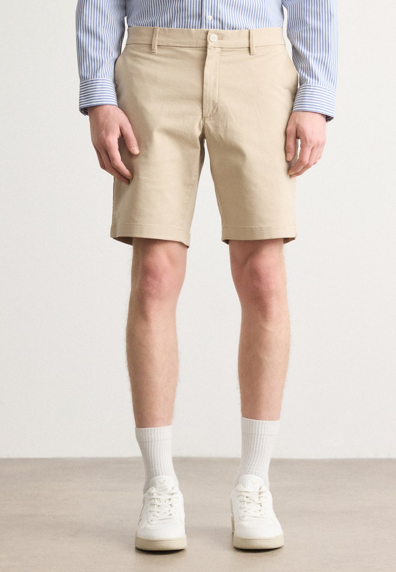 Beige bomullsshorts med brettede ermer, knapp- og glidelås-lukking, og sidelommer, kombinert med hvite sneakers og ribbede sokker.