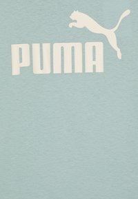 Camiseta de algodón azul claro con un gran logo blanco de "PUMA" y un gráfico de un puma saltando encima. Textura suave con un fondo liso.