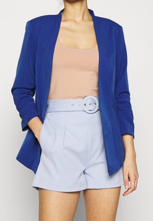 Vrouw draagt een blauwe blazer, beige tanktop en lichtblauwe short met ceintuur, met haar linkerhand in de zak.