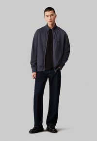 Dunkelgrau ZIP-Jacke mit hohem Kragen, getragen über einem schwarzen Hemd; kombiniert mit dunkelblauen Jeans und schwarzen Schuhen. Glatte Textur, lässiges Design.