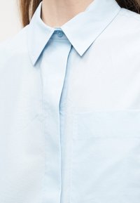 Φωτεινό γαλάζιο πουκάμισο με γιακά και κρυφή πατιλέτα κουμπιών, και τσέπη στο στήθος, φορεμένο από ένα άτομο με μακριά καστανά μαλλιά.