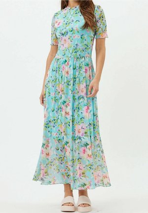 Frau trägt ein hellblaues Maxikleid mit Blumenmuster, kurzen Ärmeln und beigefarbenen Slip-on-Plattform-Sandalen vor einem einfarbigen Hintergrund.