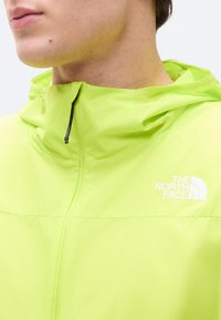 Κοντινό πλάνο ενός ατόμου που φοράει μπουφάν North Face σε φωτεινό πράσινο χρώμα, με φερμουάρ στον γιακά και εμφανές κουκούλι.