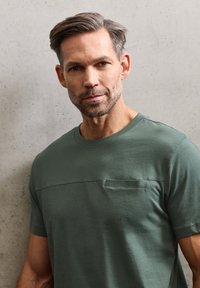 Zelené tričko s krátkými rukávy z měkké tkaniny, s náprsní kapsou, kulatým výstřihem a hladkou texturou na jednoduchém pozadí.