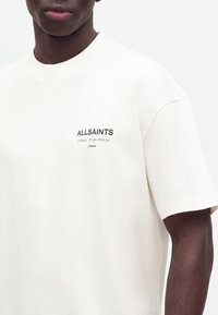 Personne à la peau foncée portant un t-shirt blanc AllSaints avec un petit texte noir sur la poitrine et des manches courtes.