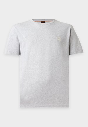 Homme debout, les mains dans les poches, portant un t-shirt gris clair, un jean ample gris foncé et des chaussures noires sur un fond blanc uni.