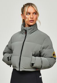 Grijze pufferjack met een hoge kraag, zwarte rits en een patchdetail op de mouw. Heeft een cropped ontwerp en elastische manchetten.