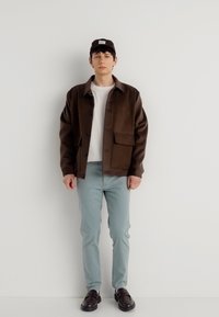 Νεαρός άνδρας που στέκεται σε έναν λευκό τοίχο φορώντας καφέ μπουφάν, λευκό πουκάμισο, ανοιχτό μπλε παντελόνι, σκούρα καφέ παπούτσια και σκούρο καπέλο.