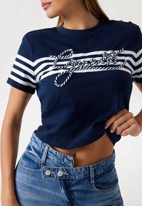 T-shirt corto blu navy con strisce bianche e dettagli in corda lungo il petto, abbinato a pantaloni corti di jeans a vita alta con dettagli a bottoni.