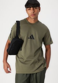 Oliven grønn kortermet t-skjorte med svart Adidas-logo; parret med en svart crossbody-veske med glidelås og justerbar reim.