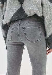 Osoba oblečená v šedých džínoch a v šedej a čiernej vzorovanej svetri, zobrazená zozadu v oblasti pásu a stehna.