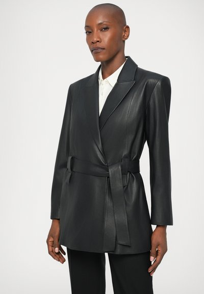 Čierny kožený blejzer s vykrojeným golierom, dlhými rukávmi a opaskom na zaväzovanie v páse. Hladká textúra a prispôsobený strih, v kombinácii s bielou košeľou pod ním.