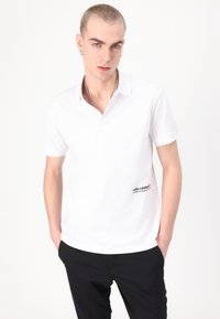 Balta trumpa polo marškinėliai, pagaminti iš medvilnės, su mažuoju juodu logotipu kairėje pusėje ir klasikiniu apykakle. Dėvimi su juodomis kelnėmis.
