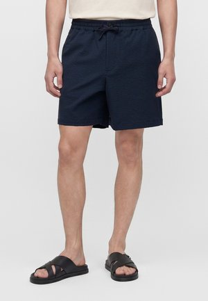 Mand iført navyblå shorts med snøre og sorte slidesandaler, stående mod en ensfarvet hvid baggrund.