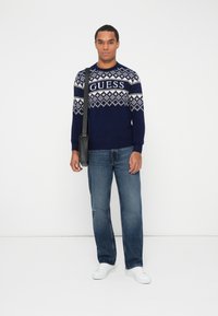 Maglione blu navy con motivo geometrico bianco e logo "GUESS", abbinato a jeans azzurri e sneakers bianche. Borsa a tracolla nera.