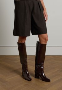 Brown pinstriped tailored shorts paired with glossy knee-high brown boots, featuring a square toe and block heel on a wooden floor.