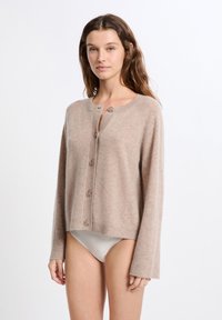 Junge Frau mit langen braunen Haaren, die einen beige geknöpften Cardigan und weiße Bikinis getragen hat, vor einem schlichten weißen Hintergrund.