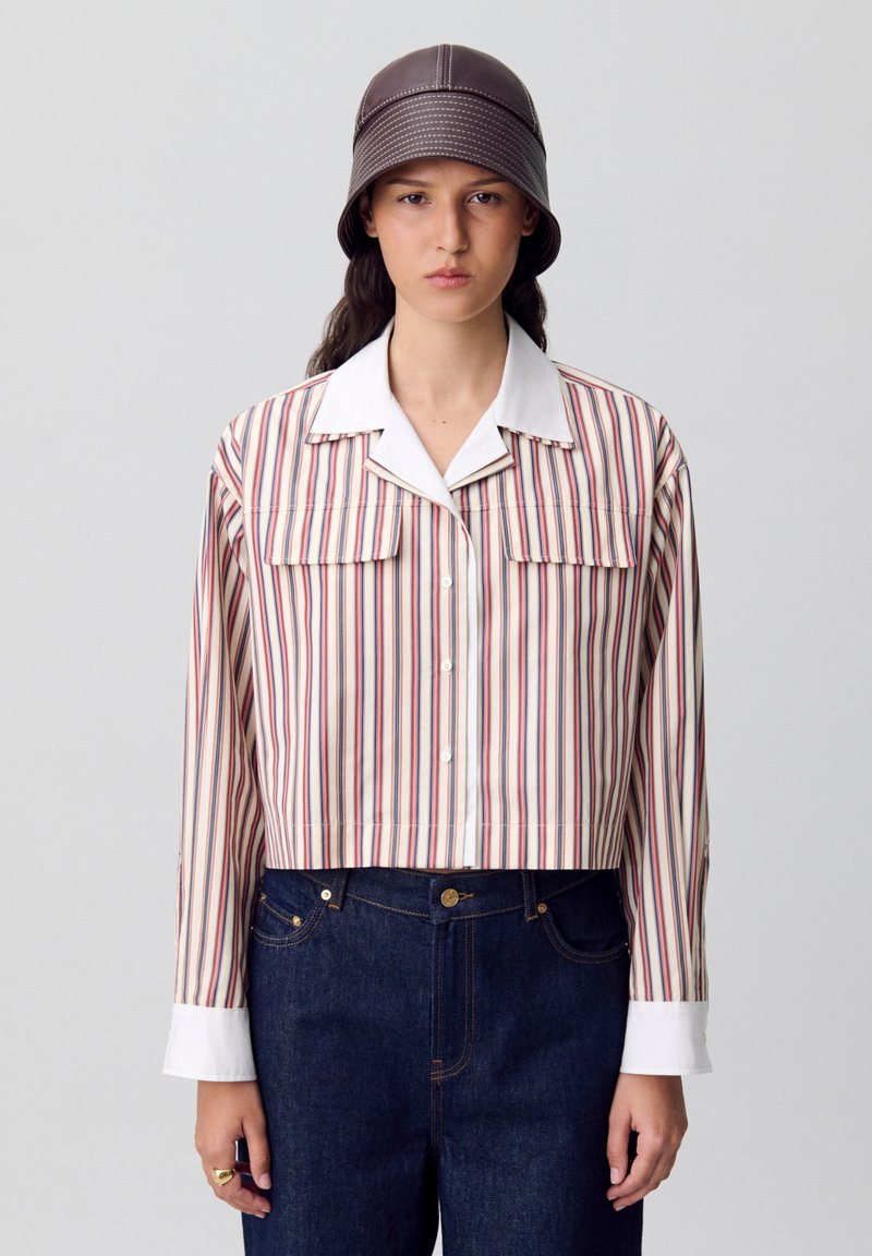 Camisa corta de botones con rayas verticales en rojo, azul y crema, con un collar blanco y dos bolsillos en el pecho. Combinada con denim oscuro.