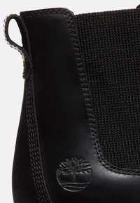 Gros plan d'une botte Chelsea en cuir noir avec panneau latéral élastique, languette de traction et logo arbre embossé près du talon.