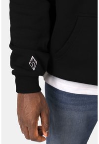 Musta huppari, jossa on ribbed hihansuut ja valkoinen timantinmuotoinen logo brodeerattu ranteen lähelle. Standardi malli, jossa on sivutasku.