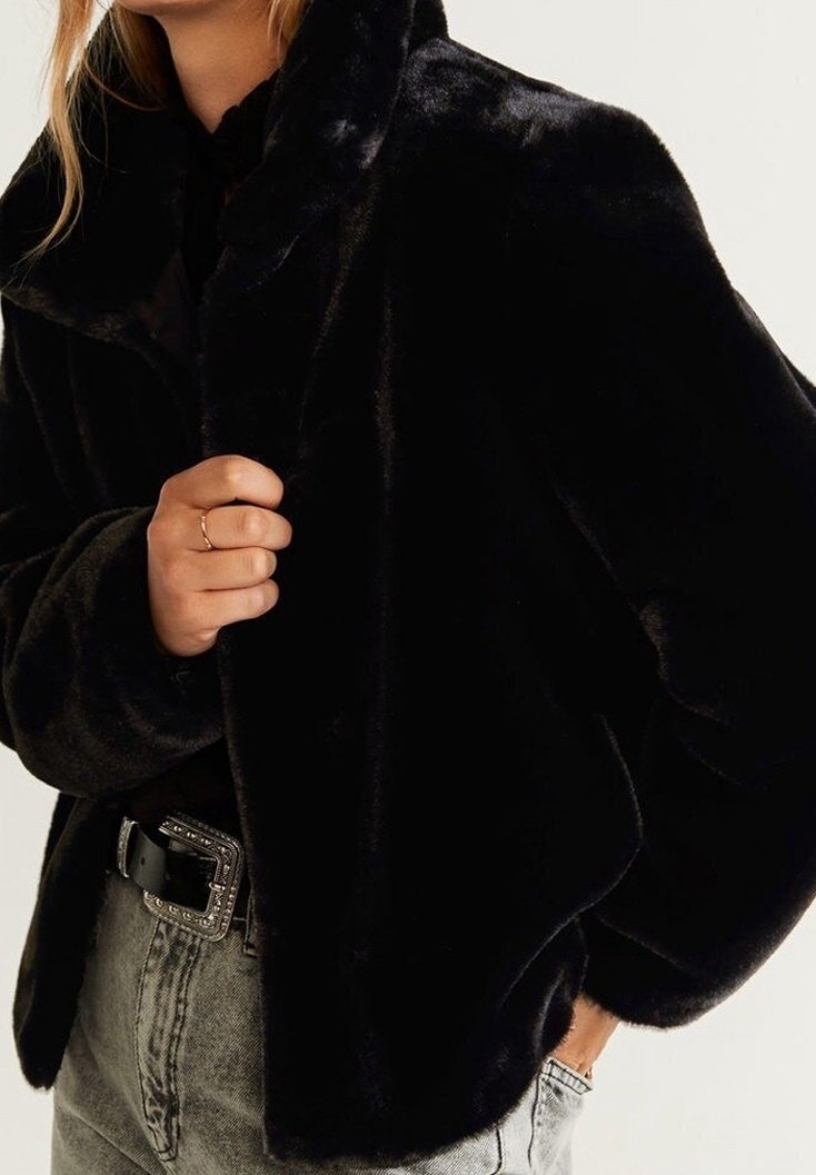 Veste en fausse fourrure noire avec un large col, une coupe oversize et une texture douce. Portée sur un jean gris clair taille haute avec une ceinture décorative.