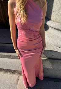 Femme portant une robe en satin rose sans manches avec des détails en dentelle, debout à l'extérieur sur des marches en pierre près d'une grande colonne en pierre.