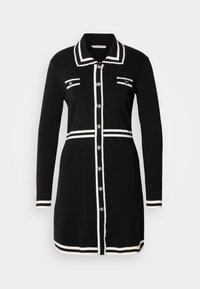 Robe noire à manches longues boutonnée avec des détails de bordure blanche sur le col, les poignets, les poches poitrine, la taille et l'ourlet.