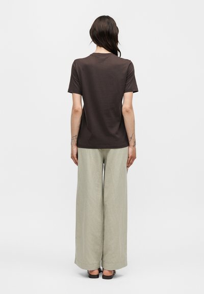 Γυναίκα που στέκεται με την πλάτη γυρισμένη, φοράει σκούρο καφέ κοντομάνικο μπλουζάκι, ανοιχτό μπεζ παντελόνι με φαρδιά μπατζάκια και μαύρα σανδάλια σε λευκό φόντο.