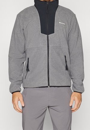 Man draagt een grijze outdoor jas met ritssluiting, zwarte kraag en ritssluiting aan de zijzakken, gecombineerd met een grijze broek.