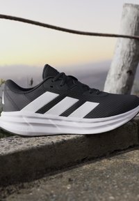 Chaussure de course noire avec des rayures blanches et une semelle amortie posée sur un rebord en pierre à l'extérieur au coucher du soleil.