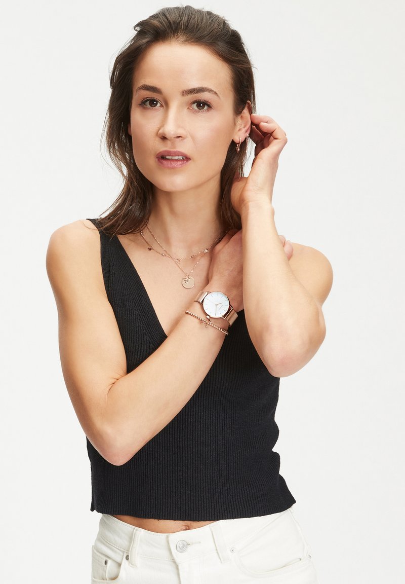 Schwarzes geripptes Crop-Top, weiße Jeans, roségoldene Uhr mit rundem Ziffernblatt, geschichtete Halsketten und Ohrringe. Neutraler Hintergrund.