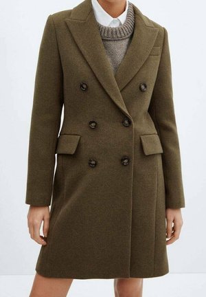 Femme portant un manteau en laine vert olive croisé sur un pull en tricot gris et une chemise blanche à col, debout avec les bras détendus.