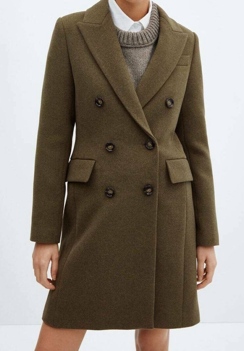 Femme portant un manteau en laine vert olive croisé sur un pull en tricot gris et une chemise blanche à col, debout avec les bras détendus.