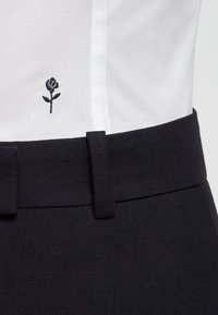 Camisa blanca con un pequeño bordado de rosa negra, metida dentro de pantalones negros de cintura alta con un lazo de cinturón ancho visible.