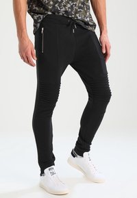 Svarta joggerbyxor med ribbade knädetaljer, sidofickor med dragkedja och en tapered passform. Tillverkade av mjukt tyg för komfort och flexibilitet.