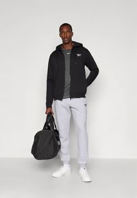 Homem em pé vestindo hoodie preto com fecho, t-shirt cinza, calças de fato de treino cinza claro, sapatilhas brancas, segurando uma mochila de ginásio preta contra um fundo simples.