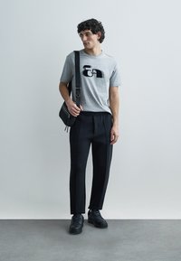 Grå bomullst-shirt med en svart grafisk logotyp, kombinerad med svarta avsmalnande byxor och svarta sneakers. Medföljer en axelväska.